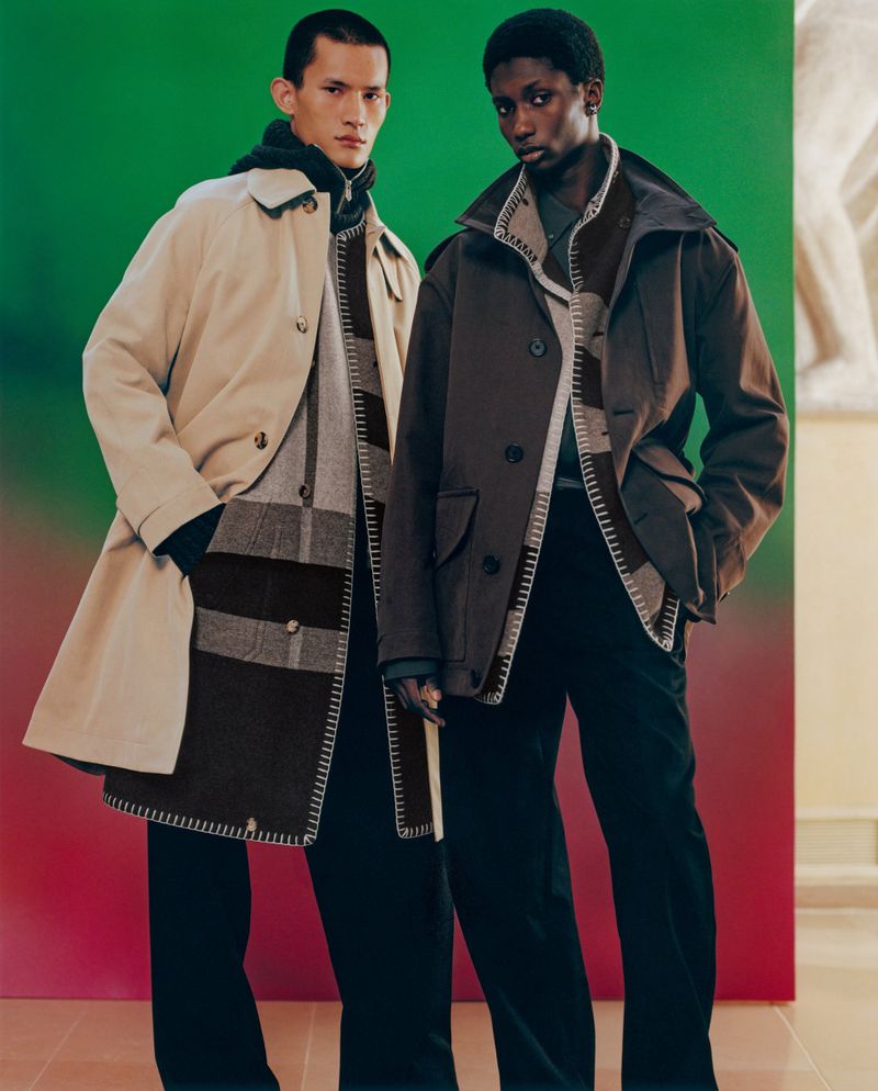 Two adult male fashion models pose indoors against a gradient backdrop wearing neutral-toned coats and dark trousers in a studio setting.