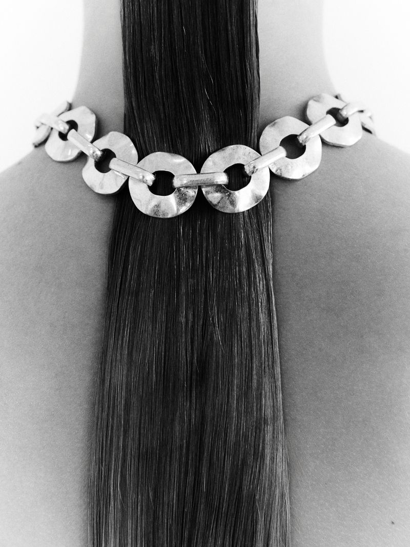 Close-up of a woman's back featuring a metal ring necklace draped over her bare shoulders, highlighting the contrast with her straight dark hair.