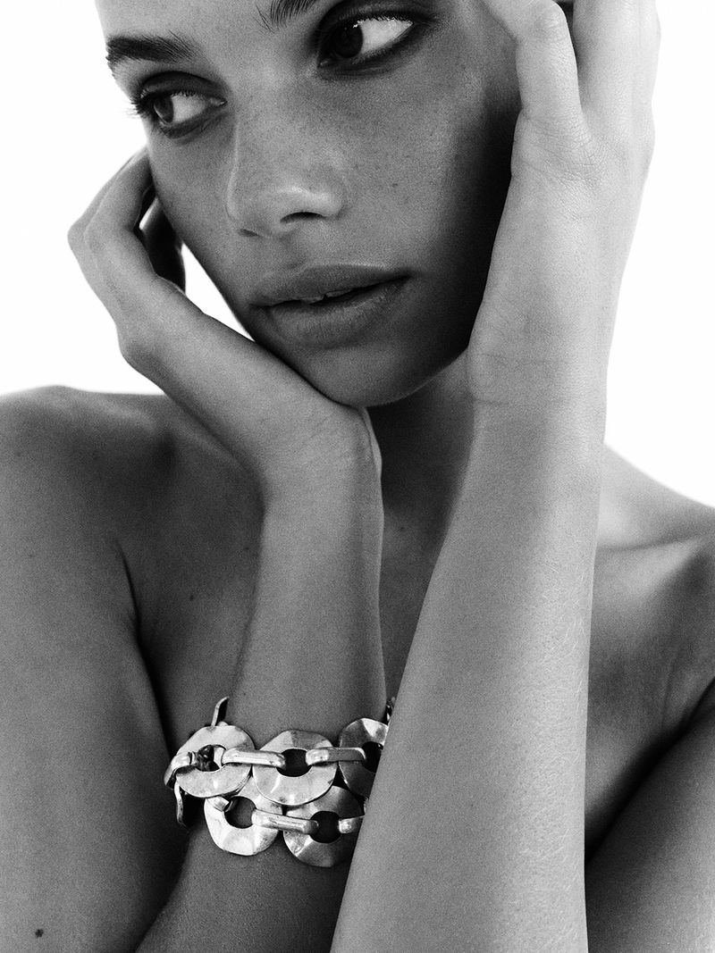 Black and white portrait of a woman with dark hair and expressive eyes, wearing a chain-link bracelet, resting her chin on her hands.