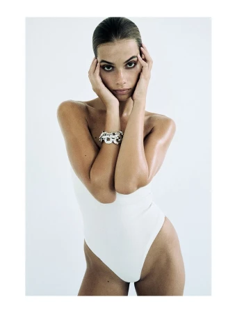 Portrait of a tanned adult female in a white bodysuit, posing with hands on her face against a bright white backdrop in a photo studio.