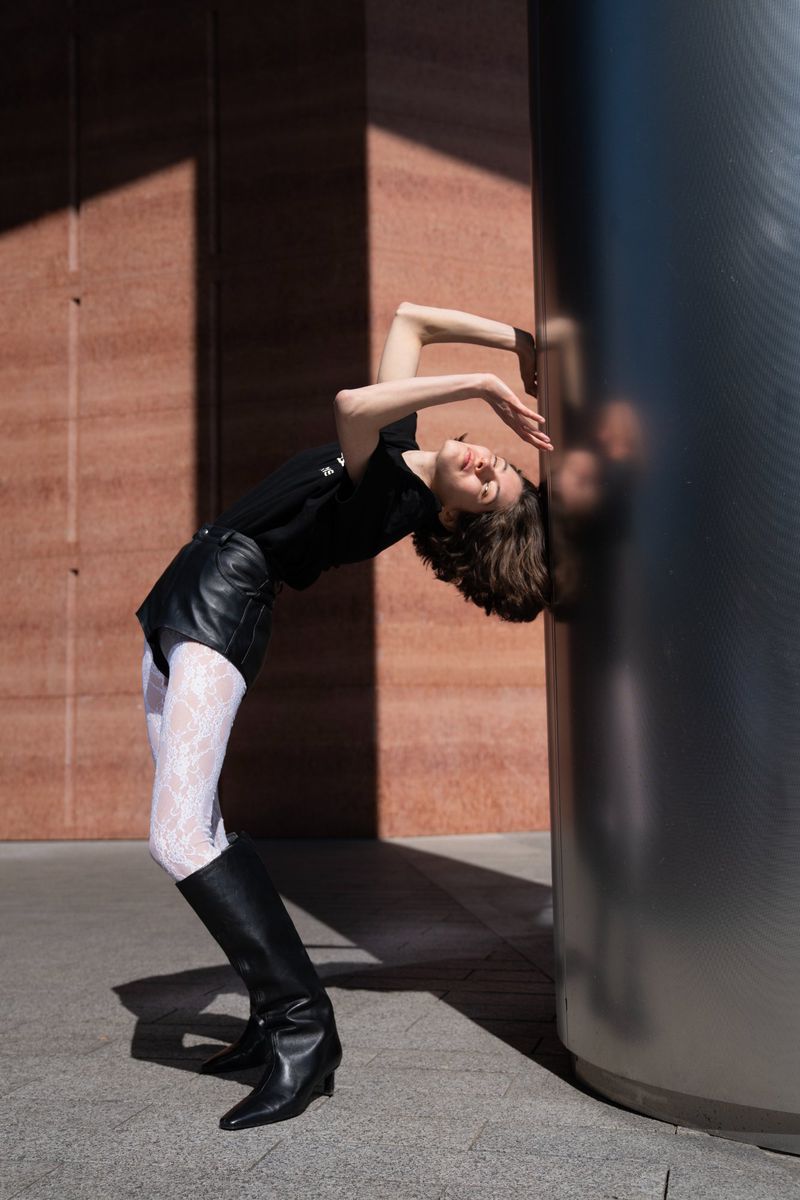 Woman in black top, leather shorts, tights and boots leans back against silver column outdoors on sunny day with shadowed wall.