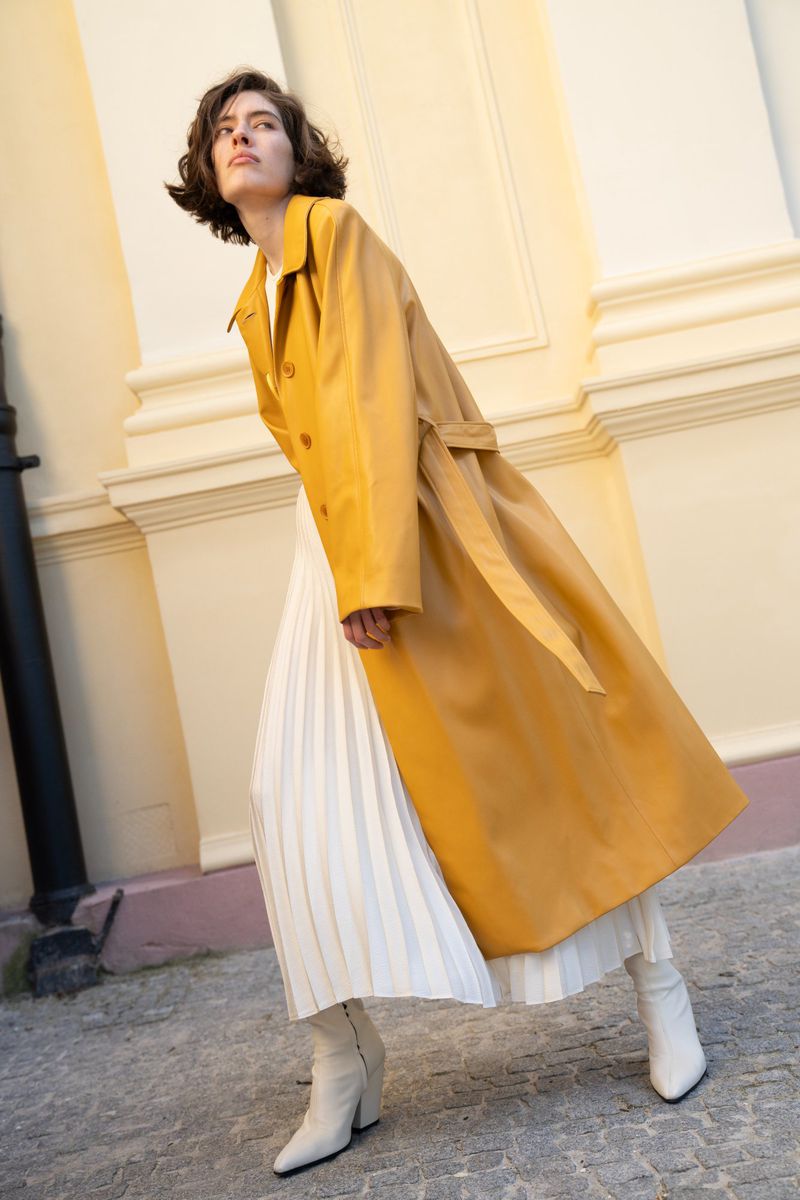 Woman in yellow trench coat and white pleated skirt walks on gray stone path in front of beige building, low angle. Fashion editorial.