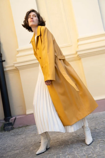 Woman in yellow trench coat and white pleated skirt walks on gray stone path in front of beige building, low angle. Fashion editorial.
