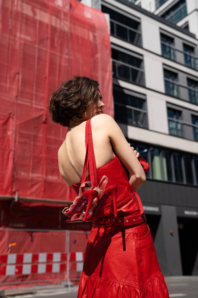 Back view of a woman in a red dress holding red shoes on a city street, with modern buildings and construction in the background.