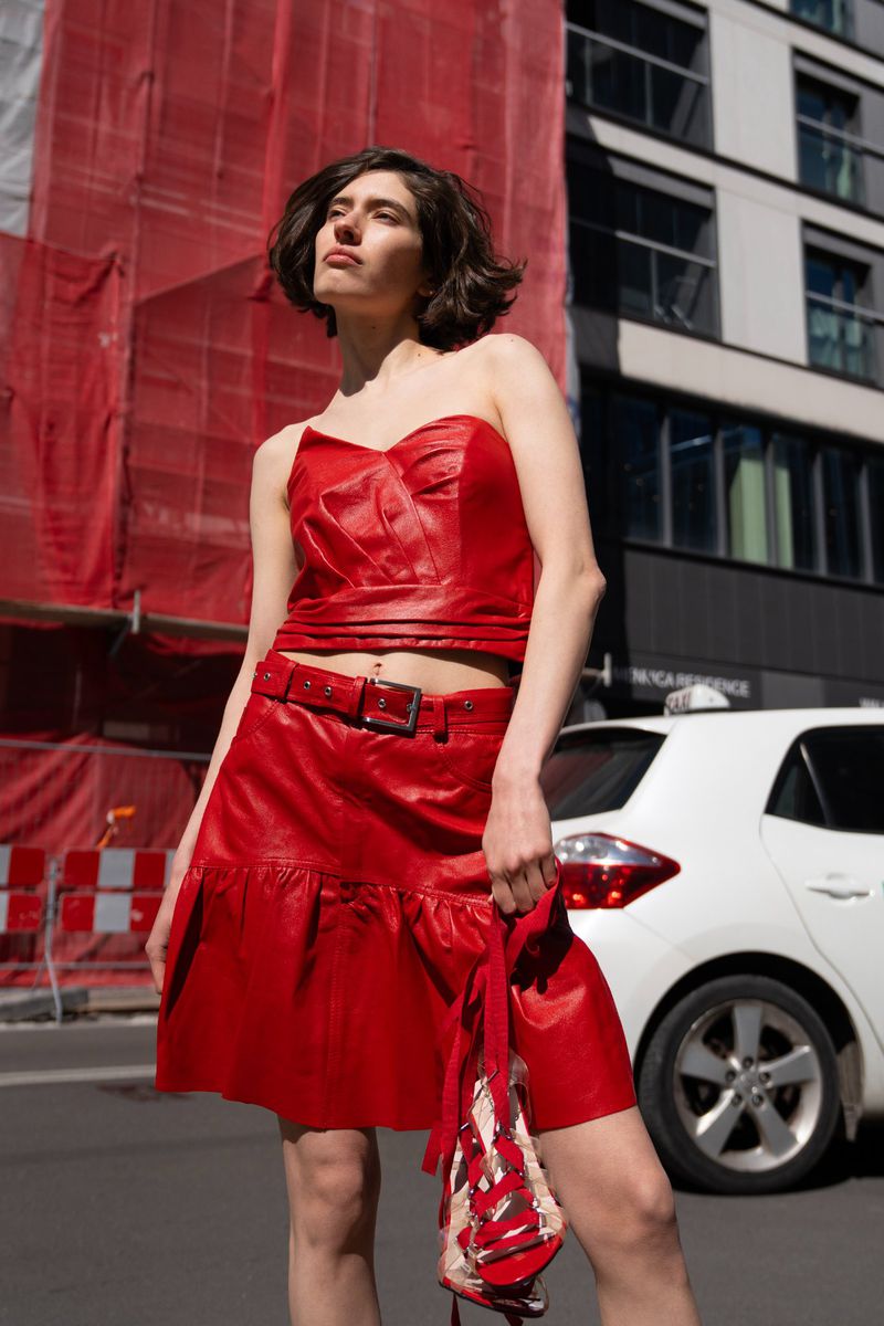 Woman in red leather two-piece outfit stands on a city street in sunlight, holding shoes, with scaffolding and a car in the background.