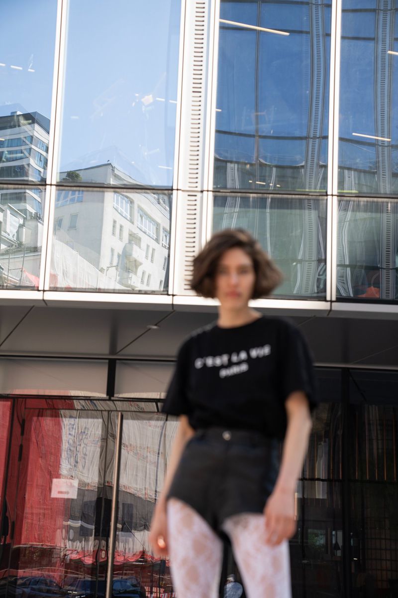 Woman in a black t-shirt with text stands in front of a modern office building with glass windows reflecting the sky.