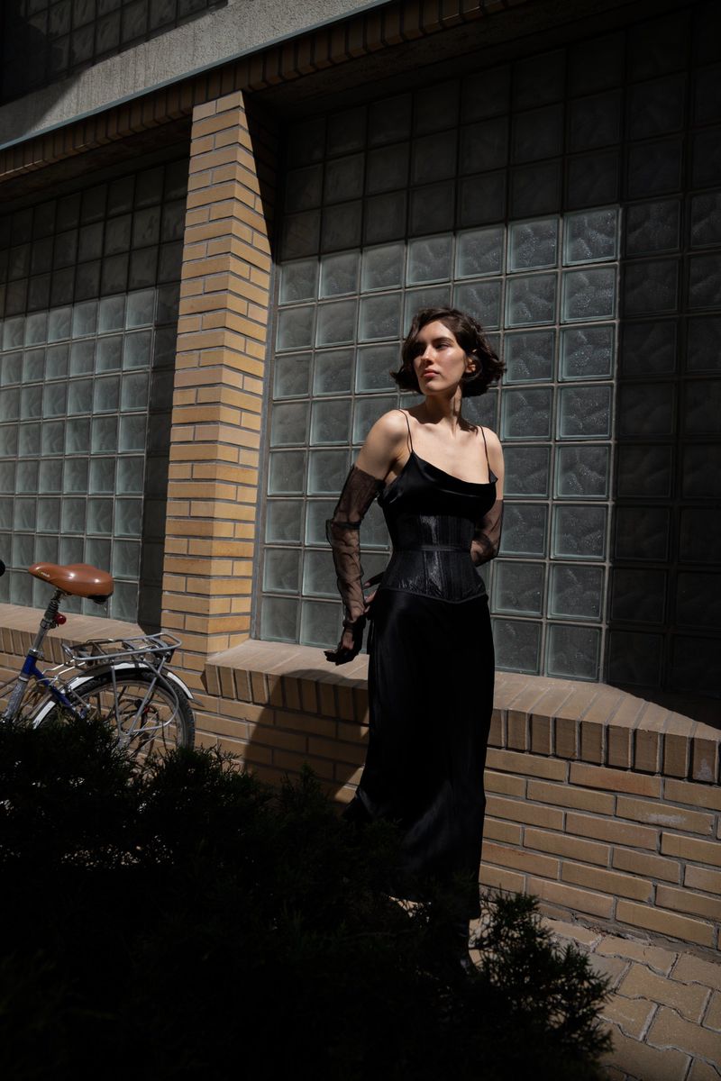 Woman in black dress poses with vintage bicycle near a brick wall with glass block windows, partially shaded by a tree.
