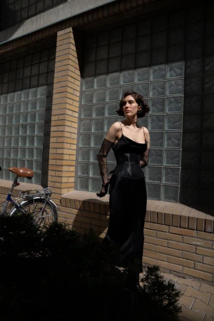 Woman in black dress poses with vintage bicycle near a brick wall with glass block windows, partially shaded by a tree.