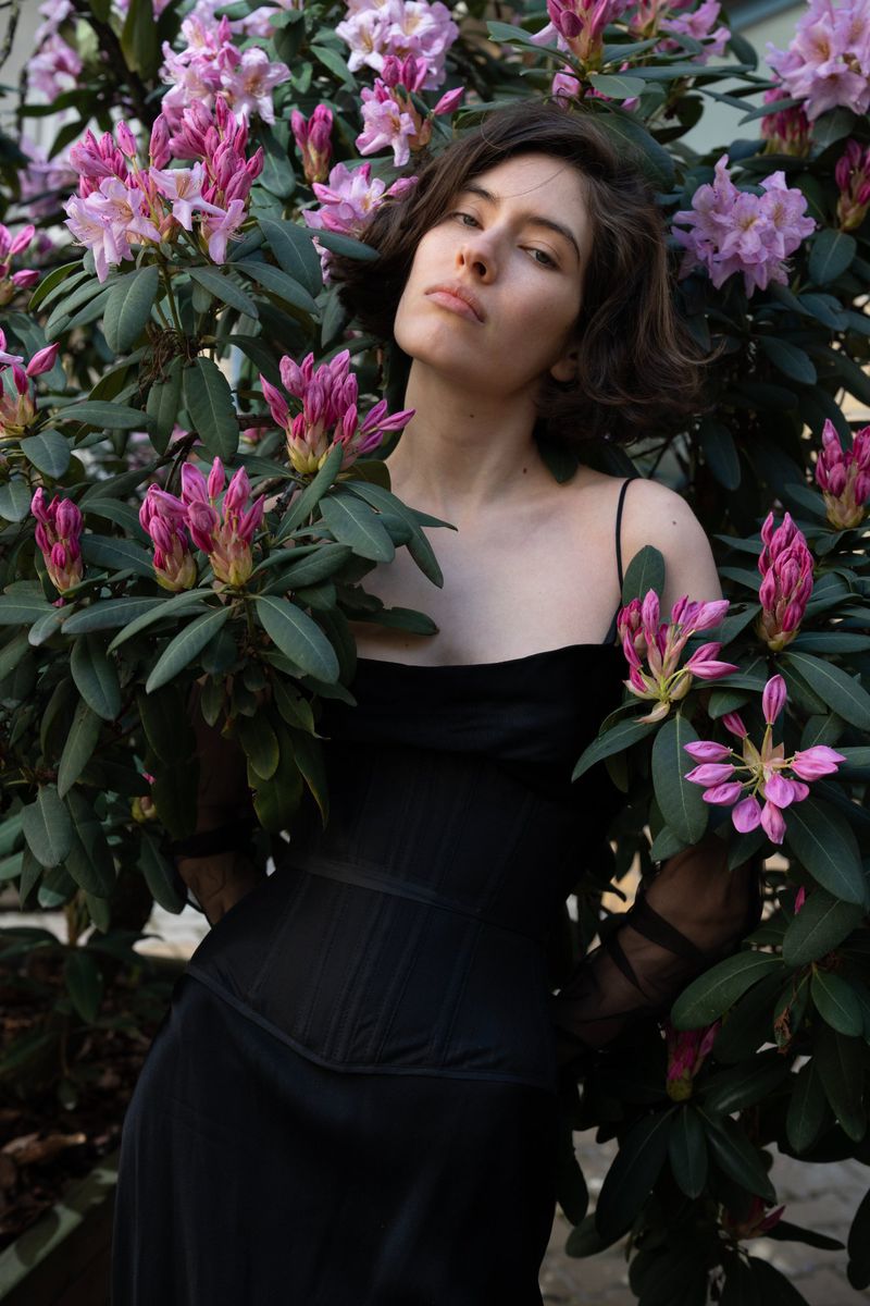Young woman in black dress poses among pink rhododendron flowers in a natural setting. Fashionable and serene outdoor portrait.