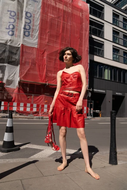 Woman in a red leather outfit stands barefoot on a sidewalk, holding red high heels, with a construction site in the background on a sunny day.