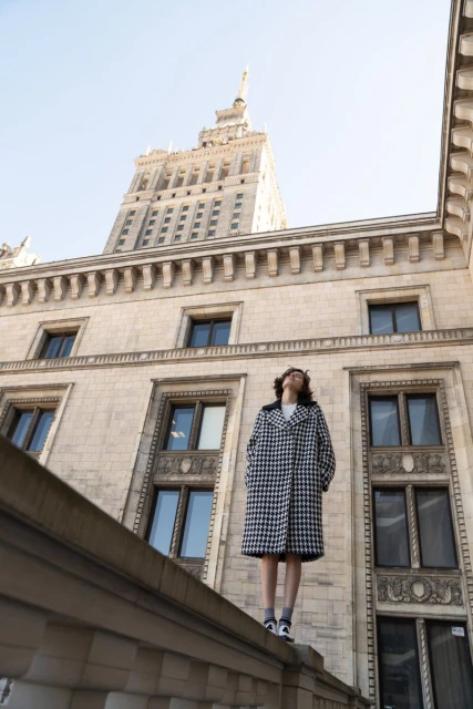 A woman in a houndstooth coat stands on a ledge against the backdrop of a tall, ornate building under a clear, blue sky.