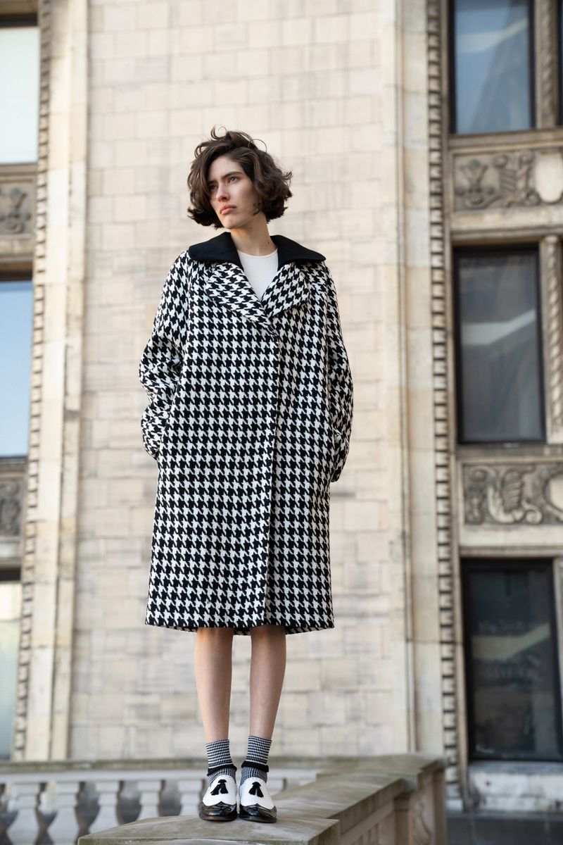 A woman in a black and white houndstooth coat stands on a ledge in front of a stone building with windows on a sunny day.