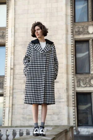 A woman in a black and white houndstooth coat stands on a ledge in front of a stone building with windows on a sunny day.