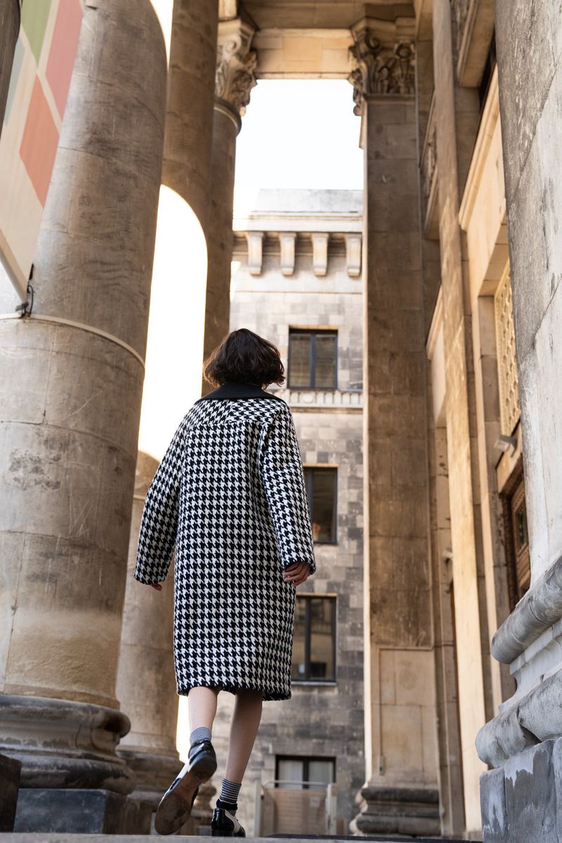Woman in houndstooth coat walks up stone steps in an urban setting with large stone columns, looking away, fashion editorial style.