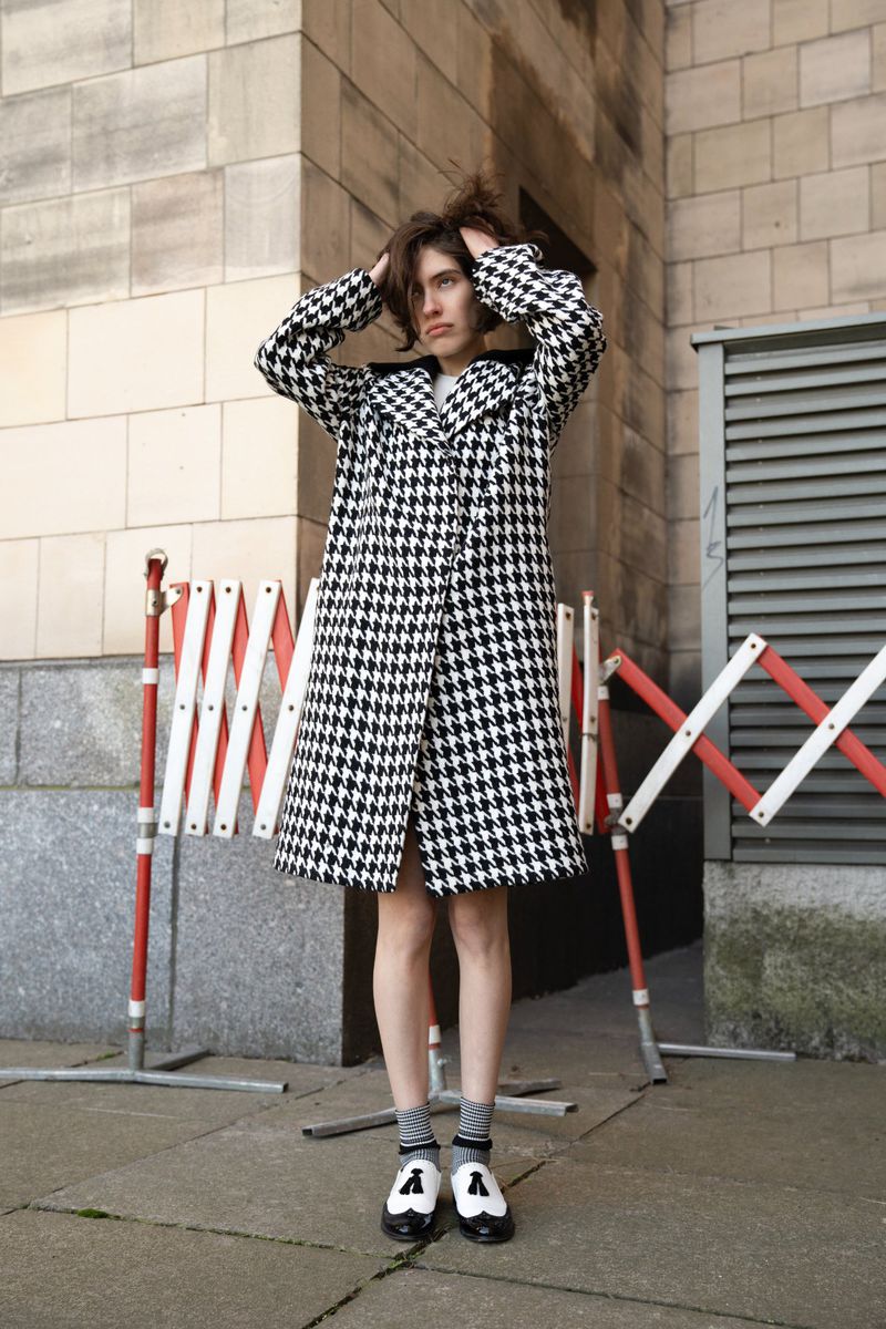 A young adult female poses on a city sidewalk wearing a black and white houndstooth coat, gray socks, and black and white saddle shoes.