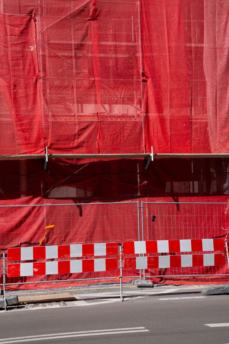 Construction site with red mesh netting covering scaffolding and a barricade with red and white squares on the roadside.