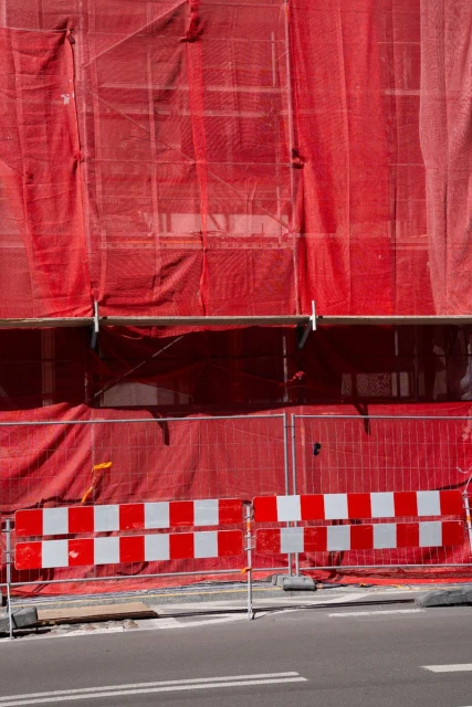 Construction site with red mesh netting covering scaffolding and a barricade with red and white squares on the roadside.
