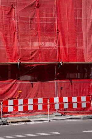 Construction site with red mesh netting covering scaffolding and a barricade with red and white squares on the roadside.