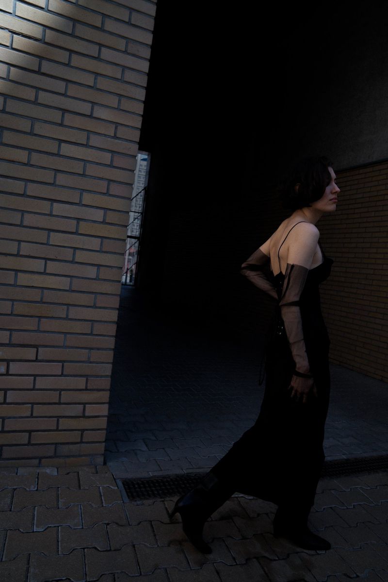 Woman in a long black dress and gloves walks along a city street beside a brick wall, dark mood, fashion photography.