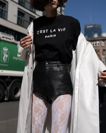 Woman in black T-shirt with 'C'est La Vie Paris' text, black leather shorts, and white patterned stockings outdoors on a sunny day.