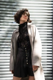Woman in dark lace top and high-waisted shorts wearing light grey coat against slatted background