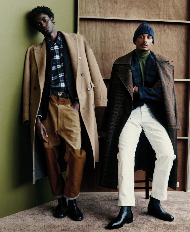 Two dark-skinned men pose in overcoats, one in tan with tan/brown pants, the other in a brown plaid coat, wearing a navy beanie and white pants.