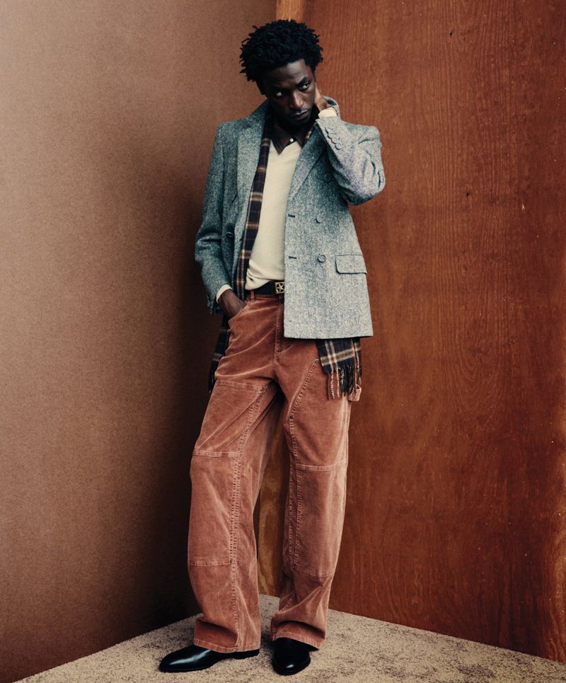 Full-length portrait of a Black man in an tweed blazer, collared shirt, corduroy pants, and leather shoes, posing against a corner.