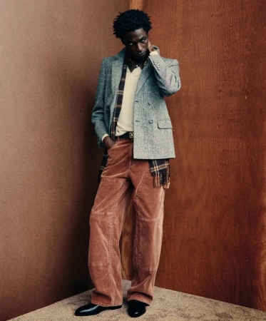 Full-length portrait of a Black man in an tweed blazer, collared shirt, corduroy pants, and leather shoes, posing against a corner.