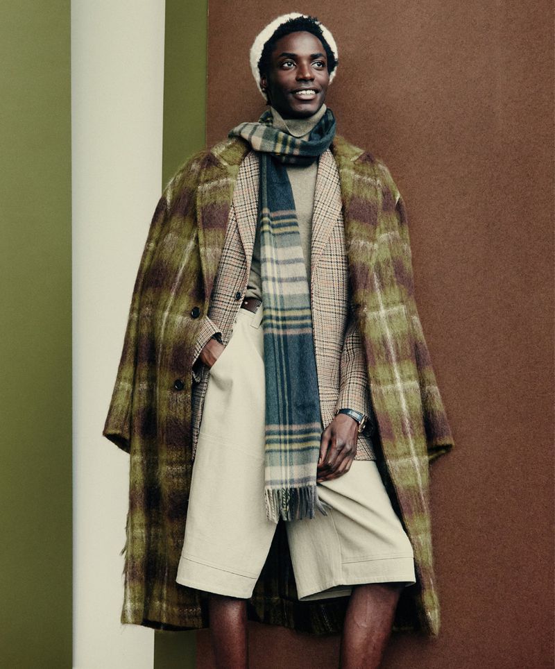 Male fashion model in a plaid coat, scarf, and beanie poses in front of a neutral backdrop with modern style and neutral colors.