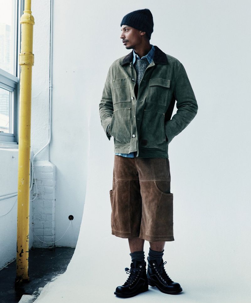 Man in olive green jacket, brown shorts, and black boots posing in a studio with a large window and white background for fashion editorial.