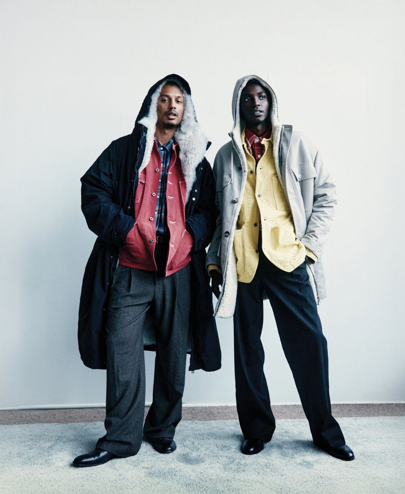 Two Black male fashion models pose indoors against a white wall. The men wear winter coats, long pants, and black shoes.
