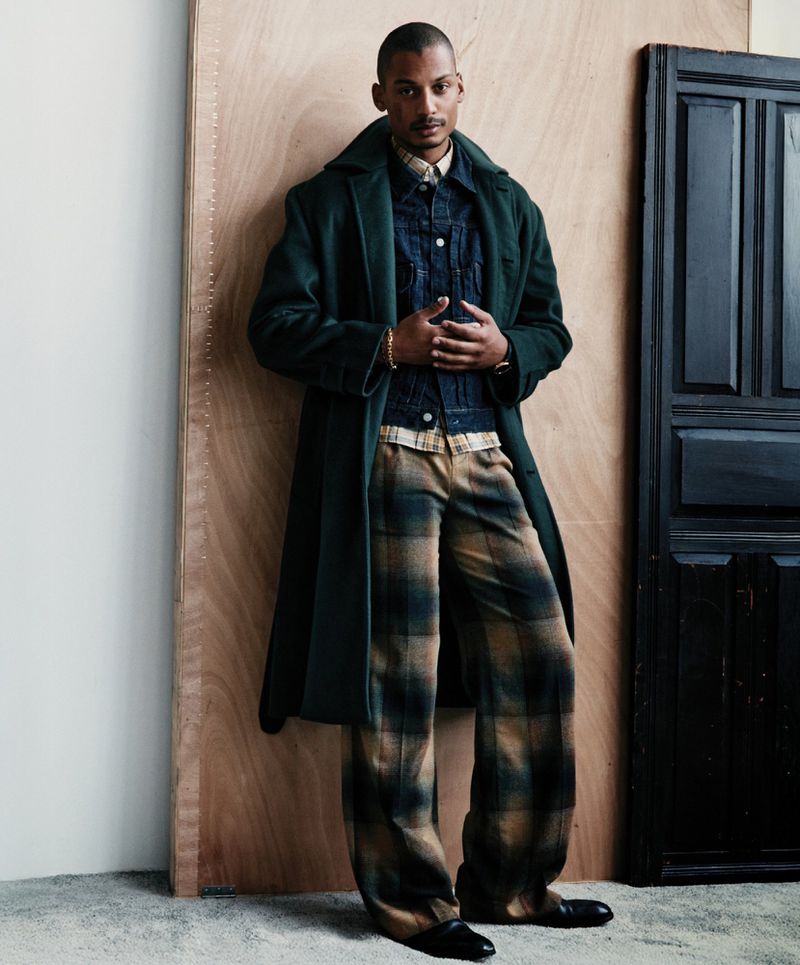 A stylish adult male with dark hair poses in an indoor photo studio, wearing a long green coat and plaid pants, against a wood background.