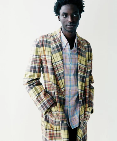 A Black male model wearing an oversized yellow and brown plaid coat over a pastel check shirt against a white seamless backdrop.
