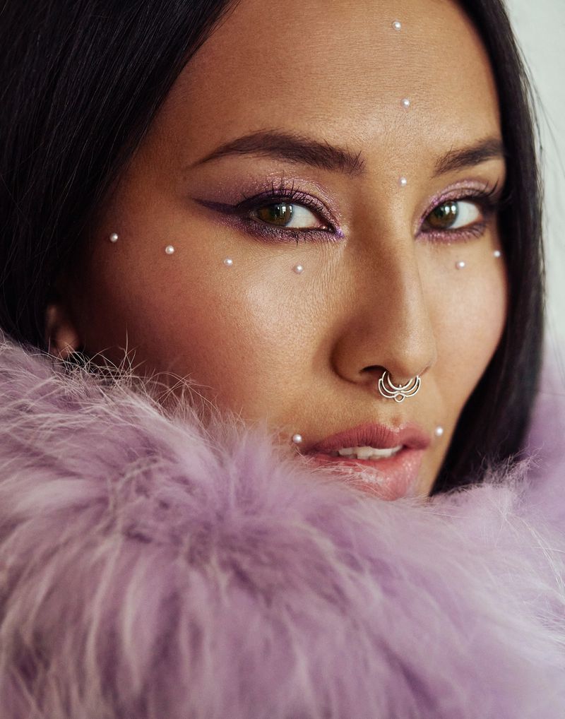 Close-up of a young adult woman with pearls on her face, dramatic eye makeup, and a nose ring, wearing a light purple feather boa.
