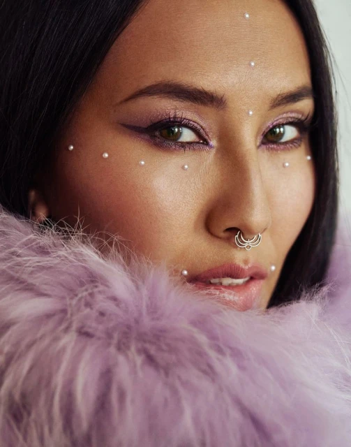 Close-up of a young adult woman with pearls on her face, dramatic eye makeup, and a nose ring, wearing a light purple feather boa.