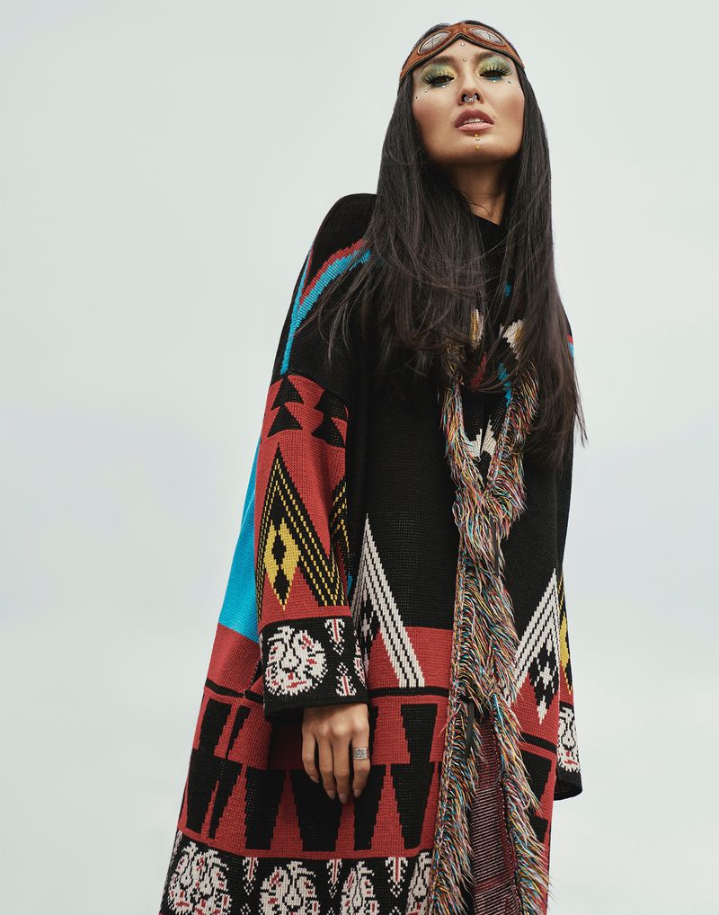 Woman wearing a patterned black coat with colorful geometric designs and fringe, and an aviator-style headband, against a bright sky.
