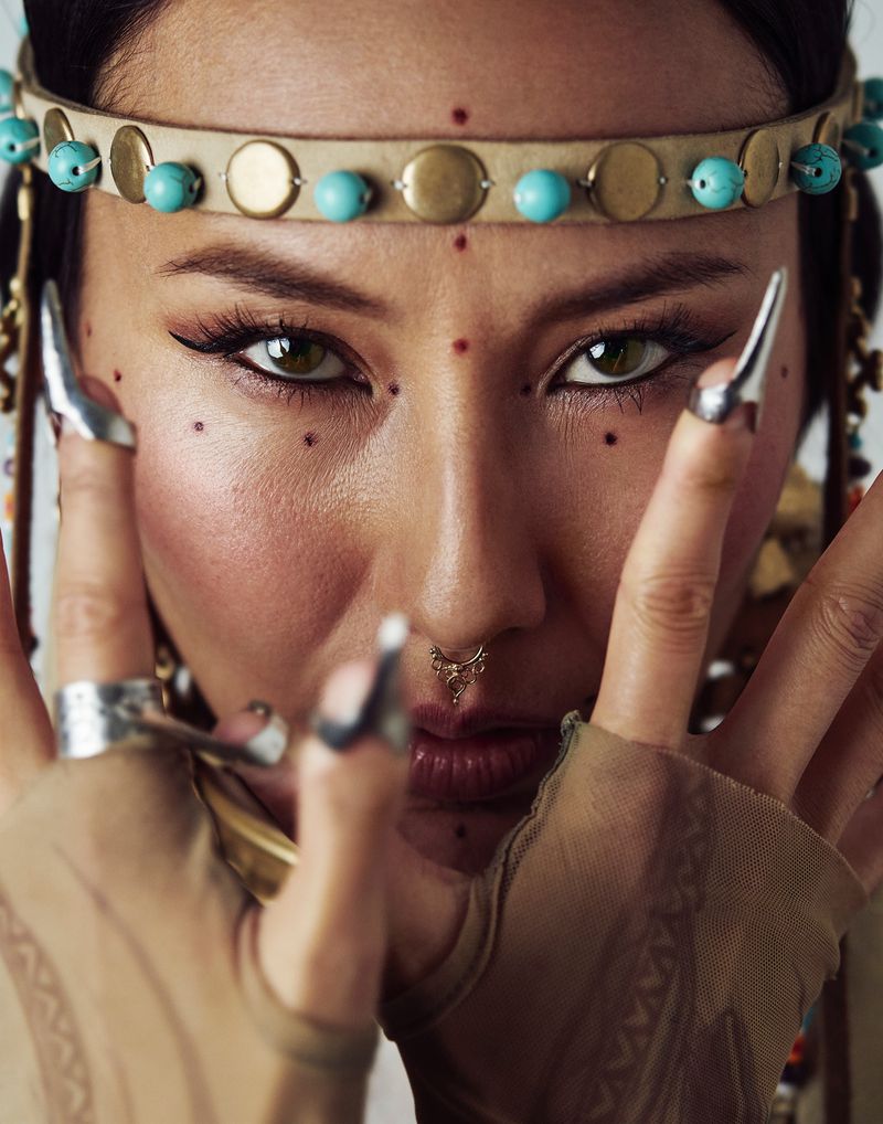 Close-up portrait of a woman with a beaded headband and decorative face dots, accessorized with large silver rings and claw-like nail extensions.