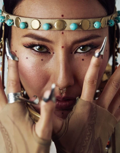 Close-up portrait of a woman with a beaded headband and decorative face dots, accessorized with large silver rings and claw-like nail extensions.