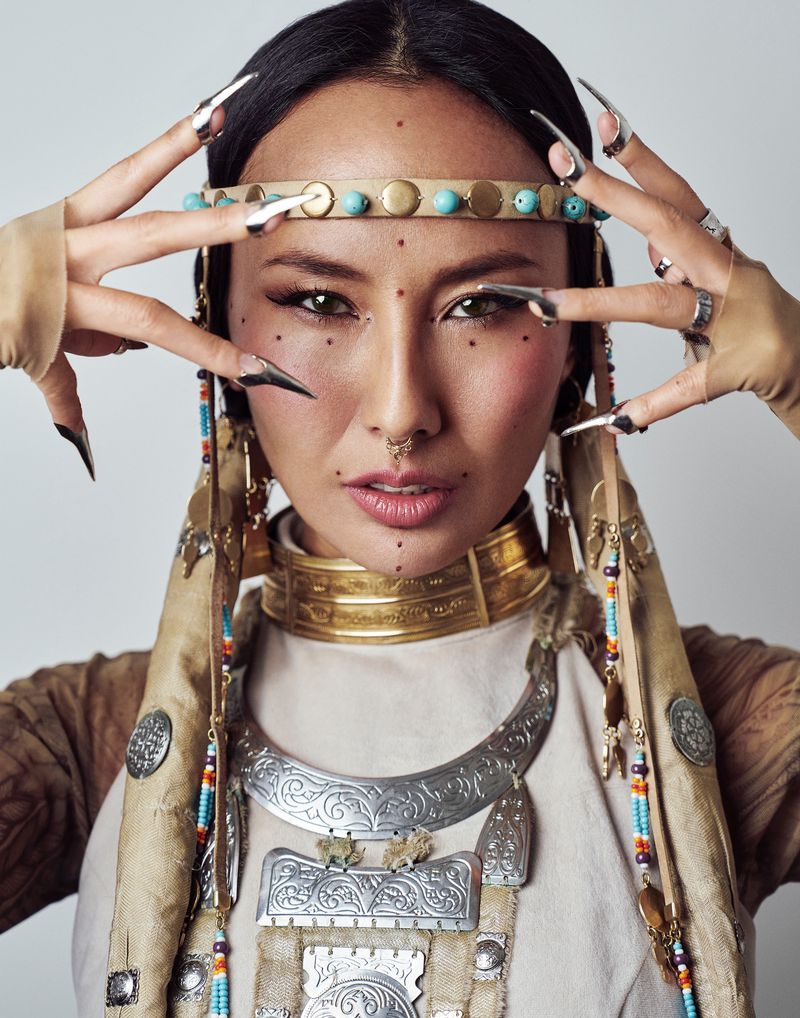 Portrait of an adult woman with long dark hair and tribal-style jewelry posing in front of a solid white background in a photo studio.