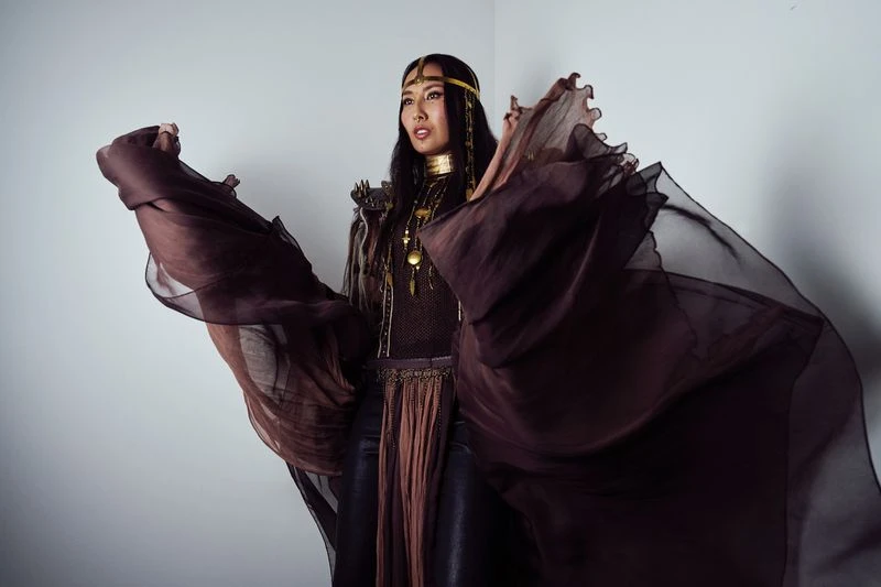 An Asian woman poses in dramatic brown chiffon fabric, wearing leather pants, gold collar and headpiece against a plain background.
