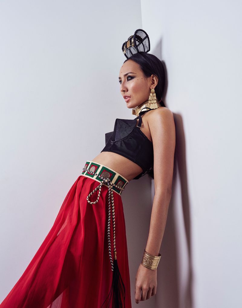 Elegant woman in black crop top and flowing red skirt, adorned with gold accessories, leans against a corner in a fashion forward pose.