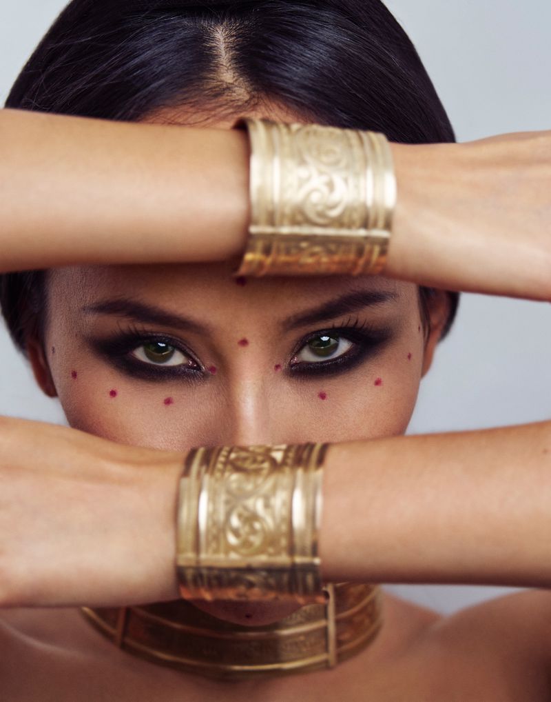 Close-up portrait of a woman with dramatic eye makeup, wearing gold cuffs and a choker, with red dots on her face against a gray backdrop.