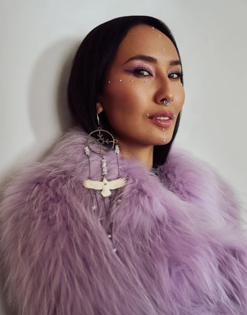 Portrait of an adult woman with dark hair, wearing purple eyeshadow and fur coat, adorned with face jewels, and custom jewelry.
