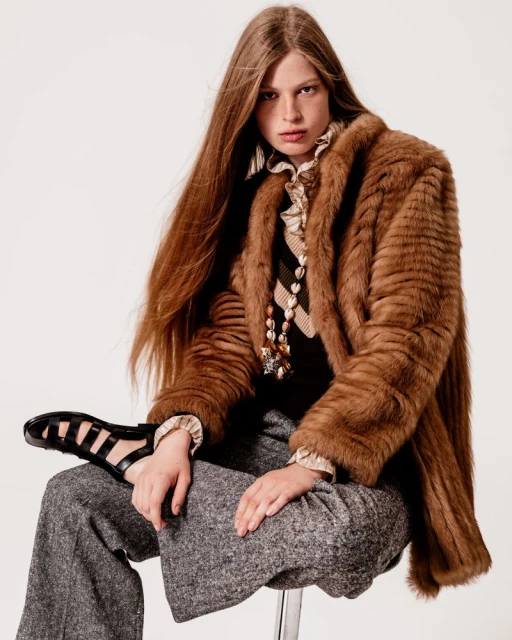 A person with long brown hair, wearing a fur coat and grey tweed pants, sits on a stool against a white backdrop in a fashion shot.