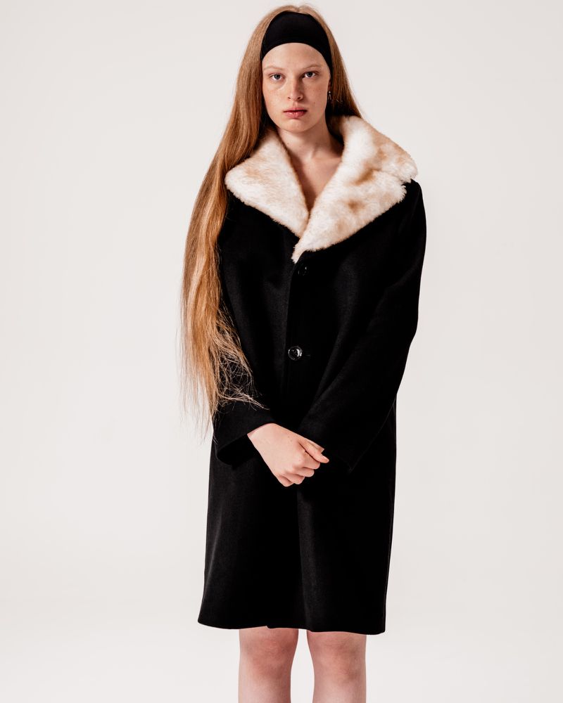 A woman with long hair wearing a black coat with a faux fur collar poses in front of a white background in a studio shot.