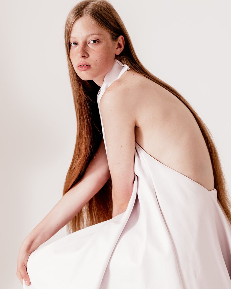 Young woman with long red hair, draped in a white fabric, sits in a bright studio, back partially visible in a fashion portrait.