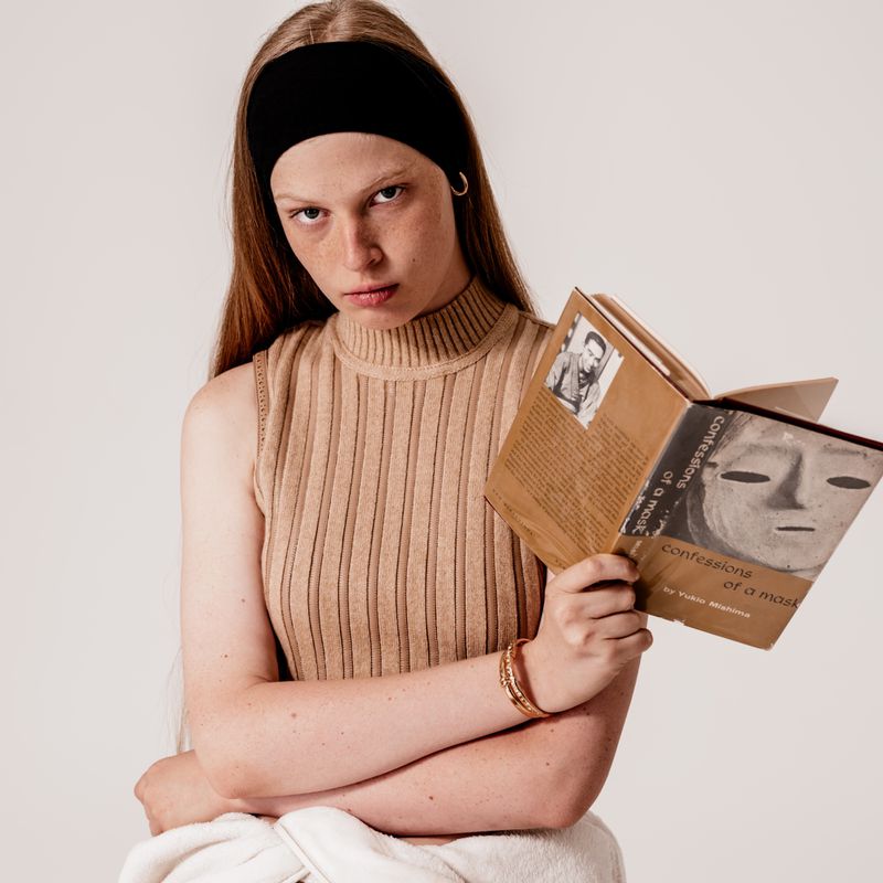 Woman reading a book with a mask on the cover, wearing a black headband and neutral-tone ribbed top, portrait in front of white backdrop.