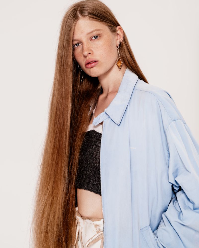 Vivian Wilson with waist-length straight hair wearing an oversized light blue jacket and dangling earring against a white background.