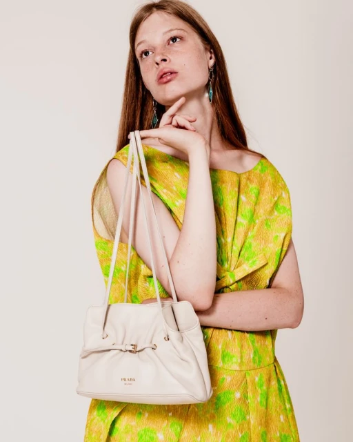 A poised woman showcases a cream-colored leather handbag, styled with a yellow and green dress and turquoise earrings in a studio setting.