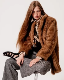 Woman with long brown hair wearing a brown fur jacket, patterned blouse, and grey trousers against a white background